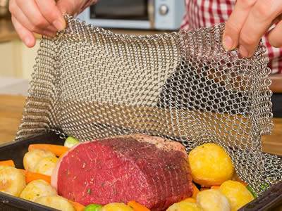 A man is covering the ss chainmail baking cover onto ovenware filled with meat and fruit.